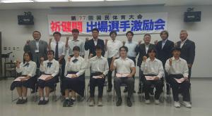 「第77回国民体育大会 祈健闘 出場選手激励会」と書かれた横断幕が会場後方の壁に掲げられ、その前方で前列には椅子に座る学生たち7名、後列に市長を含む関係者9名が立って、全員が右の肘を曲げ握りこぶしを顔の横に掲げたポーズをとり写っている記念写真