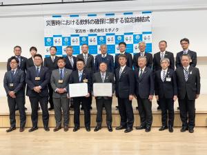 「災害時における飲料の確保に関する協定締結式」と書かれた横断幕が貼られたバックパネルの前で、前列に締結書を広げて立つ市長と男性を中心に7名の男性、1段上がった後列に10名の男性たちが立って横一列に並んでいる写真