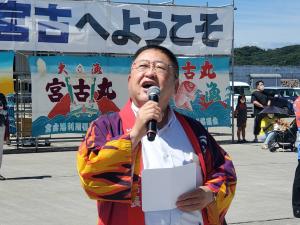 「宮古へようこそ」と書かれた横断幕や大漁旗が設置されている前で法被を着てマイクをもち大きく口を開け話をしている様子の市長の写真