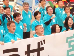 市長を中心に看板を持った職員の方達が前列、2列目以降には肘を曲げ握った拳を顔の横に掲げてポーズをとる職員達が順に並んでいる場面を写した写真