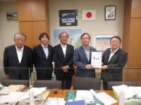 中央に透明のパーテーションが設置されたテーブルの傍で、自由民主党の谷公一衆議院議員と市長が要望書を一緒に持ち、宮古市議会の橋本久夫議長と宮古商工会議所の大坂文人副会頭、関係者の男性が横一列に並んでいる写真