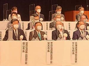白いテーブルクロスが敷かれ前面に名前札が貼られた机が階段のように3段に並び、白い胸章を着けた関係者たちがそれぞれ座っている写真