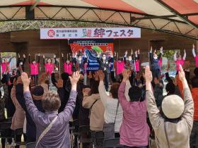 絆フェスタと書かれた横断幕の下のステージに立っている方々と参加者の方々が全線開通を祝って万歳をしている写真