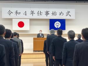 「令和4年仕事始め式」と書かれた横断幕と国旗、宮古市の市町村旗が吊るされた壇上に置かれた演台の前で話をする市長と、壇上を向いて立って市長の話を聞く職員たちの写真