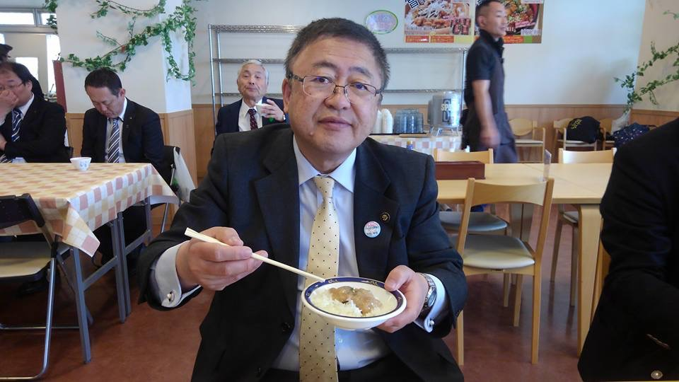 右手に割りばし、左手に丸皿を持ち、お皿に入った料理を見せている様子の市長の写真