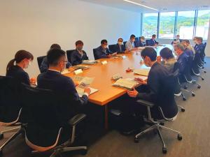 部屋の中心に置かれた木製の大きなテーブルの周りを囲むように、関係者の方たちが大勢座り、首を少し下に向け手元の資料に目を通している様子の写真