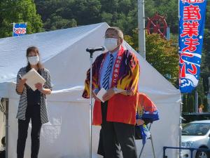 法被を着てスタンドマイクの前に立つ市長と、両手に持った資料を見ながら市長の斜め後方に立つ女性を写した写真