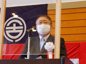 後方の壁に市旗が貼られた会場で、パーテーションが設置された檀上に白色の胸章リボンを付けた市長が立ち挨拶を行っている「国道340号宮古岩泉間整備促進住民総決起大会」の様子の写真