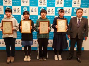 賞状の額縁を持った4名の女子学生と市長がバックパネルの前に横一列に並んでいる写真