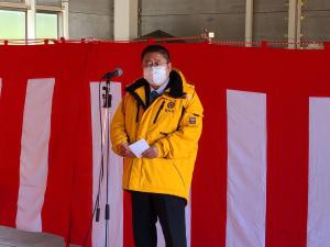 紅白幕が飾られた安全祈願祭と除雪出動式の会場で、黄色のジャンパーを着用した市長がマイクスタンドの前で挨拶を行っている様子の写真
