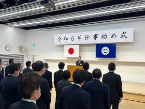 令和6年仕事初め式とかかれた幕が飾られ、並んでいる職員にむかって演台から挨拶をしている市長の写真