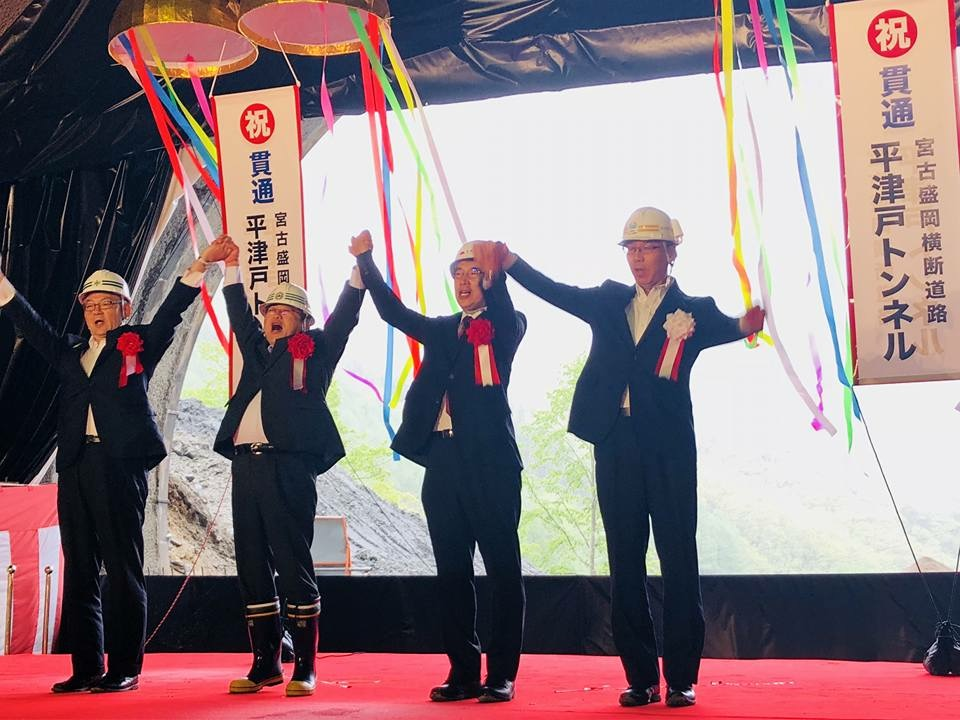 赤色、黄色、青色などの紐が垂れ下がる割れたくす玉の下で、市長を含む4名のヘルメットを被った関係者の方々が手をつなぎ両手を上に掲げている写真