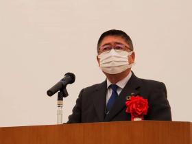 会場の演台で、赤の胸章をつけた市長があいさつをしている写真