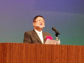 開校式の演台で赤の胸章をつけた市長が、生徒に向けて祝辞を述べている写真