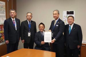市長含む関係者5名が並び、中央の車いすに座った横沢高徳議員とその横に立つ男性が要望書を持っている写真