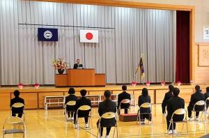 体育館のステージ上に置かれた演台で、市長が座っている制服姿の新1年生に向けて祝辞を述べている写真