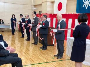 壁際に紅白幕が貼られた会場で、胸章を付け白い手袋を着けた右手にはさみを持った市長と関係者の方々4名がテープカットをしている写真