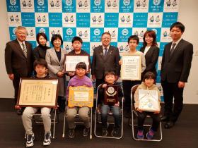 亀岳小の6人の児童たちが受賞した賞の賞状を持ち、市長、教員の方々、他の市職員の方と2列に並んで撮影した写真