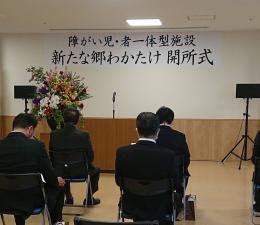 「障がい児・者一体型施設 新たな郷わかたけ 開所式」と書かれた横断幕が壁の上部に貼らた部屋の中で、椅子に座っている関係者の方たちを後方から写した写真