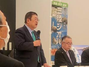「日本ジオパーク再認定にかかる首長ヒアリング」会場で、他の首長の方々が座っている席から立ち上がって右手にマイクを持ち挨拶を行っている市長の写真
