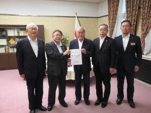 国旗が置かれた室内で、鈴木俊一財務大臣と市長が要望書を一緒に持ち、3名の関係者が横一列に並んでいる写真