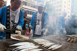 商店街で、宮古と書かれたTシャツに青いはっぴ姿の男性達が、網焼きで焼いたサンマをたくさん並べてサンマを皿にのせている写真