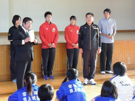 体育館の舞台前に市長と、ジャージを着た男性や女性が前に立っており、向い合わせにジャージ姿の学生が、体育座りをして話に耳を傾ける学生の写真