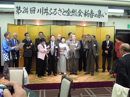 「第24回川井ふるさと会総会新春の集い」と書かれた横断幕と金の屏風が設置されたステージ上に上がっている参加者達の写真