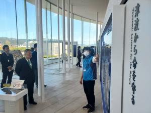 黒いスーツを着た男性たちが、津波から命を守ると書かれたパネルの内容を解説している係員の人の話を聞いている写真