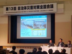 第13回自治体災害対策全国会議と書かれた幕の下で、プロジェクターで映された資料とその脇で話をする3人の写真