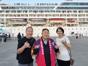 側面を見せている客船の前で、紫と赤のグラデーションの法被を着た市長とその両脇にいる外国人の3人がポーズをとっている写真