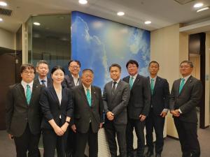 青空のような背景の前で、黒いスーツを着た市長と、スーツを着た人達が並んでいる写真