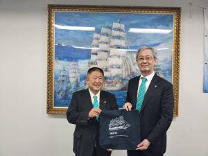 額縁に入れられた船の絵が飾られた壁の前で、スーツを着た市長と男性が船が書かれた黒い絵のようなものを、二人で並んで持っている写真