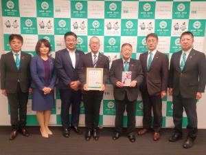 宮古市のバックパネルの前で、金封を持ったスーツを着た市長と感謝状を持った有賀義照盛岡支社長が並び、その両脇ににスーツを着た人々が並んでいる写真