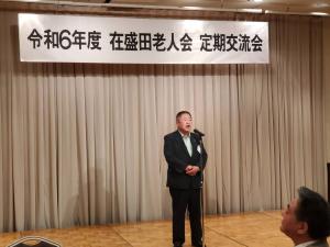 令和6年度在盛田老人会定期交流会と書かれた幕が設置されたステージの上で、マイクの前に立ち挨拶をしているスーツを着た市長の写真