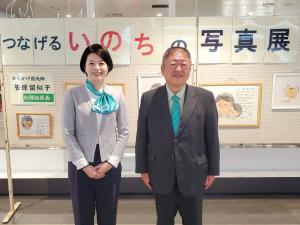 つなげるいのちの写真展と書かれたボードの前で、礼服を着た女性と黒いスーツを着た市長が並んでいる写真