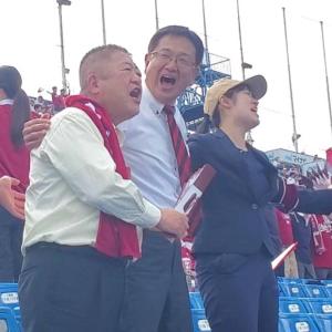 赤いタオルを首にかけレモン色のシャツを着た市長が、薄い水色のシャツを着た男性と肩を組みながら応援している写真