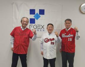 広島東洋カープのTシャツを着た市長と、2名の方が企業のロゴが入った旗が飾られた壁の前で並んでいる写真
