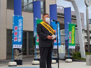 「交通安全運動実施中」と書かれた幟が4つ並べられた広場の前で黄色い襷を掛け両手で手紙を持ち話をしている様子の市長の写真