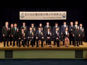「宮古地区優良勤労青年表彰式」と書かれた横断幕が舞台後方に掲げられ、前列に椅子に座った6名の方達、後列に起立して横に並ぶ12名の方達が写った記念写真