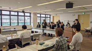 長机がロの字型に設置された会議室に参加者が着席し、前方の席から立ち上がった市長が挨拶を行っている様子の写真