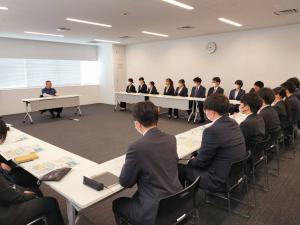 市長が窓際に置かれた長テーブルの席に着き、それを囲むように、長テーブルをいくつか並べてコの字型に置かれた席に数十名の職員が座り、市長の話を聞いている様子の写真