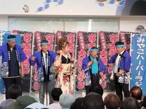 幟が立っているステージで、法被を着た市長を含む男性4名の前で、着物姿の女性がマイクを持ち話をしている写真