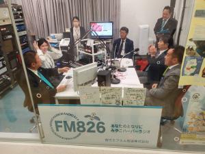 FM826と書かれたボードがクリアガラスに立てかけられた部屋で市長、男性3名、女性1名が机を囲むように座っている写真