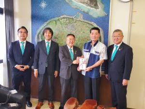 多良間島を上空から撮影した大きな写真が飾られた壁をバックに、中央に伊良皆光夫村長と市長が一緒に御祝金ののし袋を持ち、3名の関係者が横一列に並んでいる記念写真