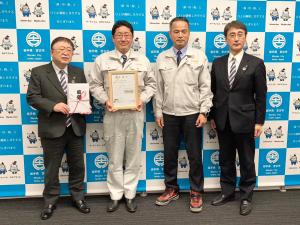 左から目録を持った市長、額縁を持った作業服の男性と関係者の男性2名がバックパネルの前で横一列に並んでいる写真