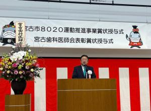 「宮古市8020運動推進事業賞状授与式 宮古歯科医師会表彰賞状授与式」と書かれた横断幕や紅白幕、カラフルな色の豪華な壇上花が飾られたステージの檀上で挨拶を行っている市長の写真