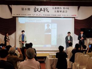 「第八期叡王戦」と書かれた看板の下に大盤解説が映されたスクリーンが置かれ、そのスクリーンの両サイドに藤井聡太叡王と菅井竜也八段が立ち、椅子に座ってスクリーンの方を見る参加者たちの写真