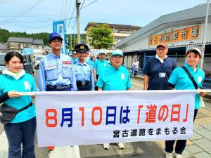 「8月10日は「道の日」 宮古道路をまもる会」と書かれた横断幕が垂らされた棒を持つ女性2人と、その後ろで市長や関係者の方々が並んでパレードをしている様子の写真