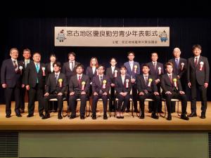 「宮古地区優良勤労青少年表彰式」と書かれた横断幕が設置されたステージ上で、左胸に胸章リボンを付けた受賞者や市長、関係者の方々が前列の椅子に6名、後列に11名立っている記念写真